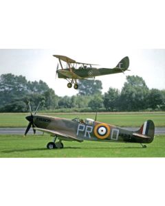 A vintage Supermarine Spitfire airplane on the ground with the registration 'AR213' and a colorful roundel, as a biplane performs a maneuver overhead against a backdrop of greenery.