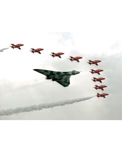 A striking aerial display featuring a vintage bomber aircraft leading a formation of red fighter jets, leaving colorful smoke trails against a cloudy sky.
