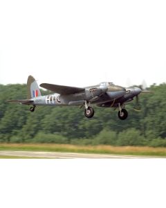 A vintage aircraft, possibly a de Havilland Mosquito, taking off against a lush green backdrop, showcasing its distinctive twin-engine design and military markings.