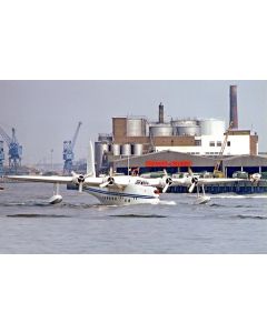 Seaplane taking off from water near an industrial harbor with storage tanks and cranes in the background.