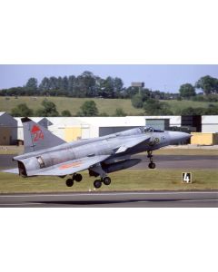 Military jet taking off from an airfield with green fields and hangars in the background.