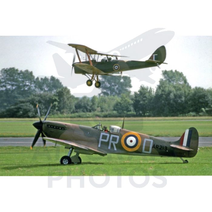 A vintage Supermarine Spitfire airplane on the ground with the registration 'AR213' and a colorful roundel, as a biplane performs a maneuver overhead against a backdrop of greenery.