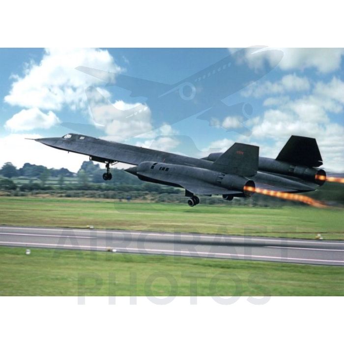 Sleek black aircraft taking off with afterburners engaged, set against a bright blue sky and green landscape.