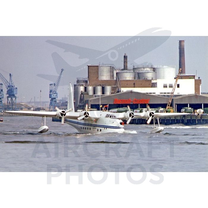 Seaplane taking off from water near an industrial harbor with storage tanks and cranes in the background.