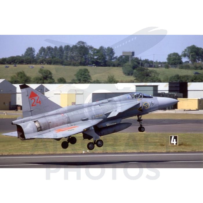 Military jet taking off from an airfield with green fields and hangars in the background.